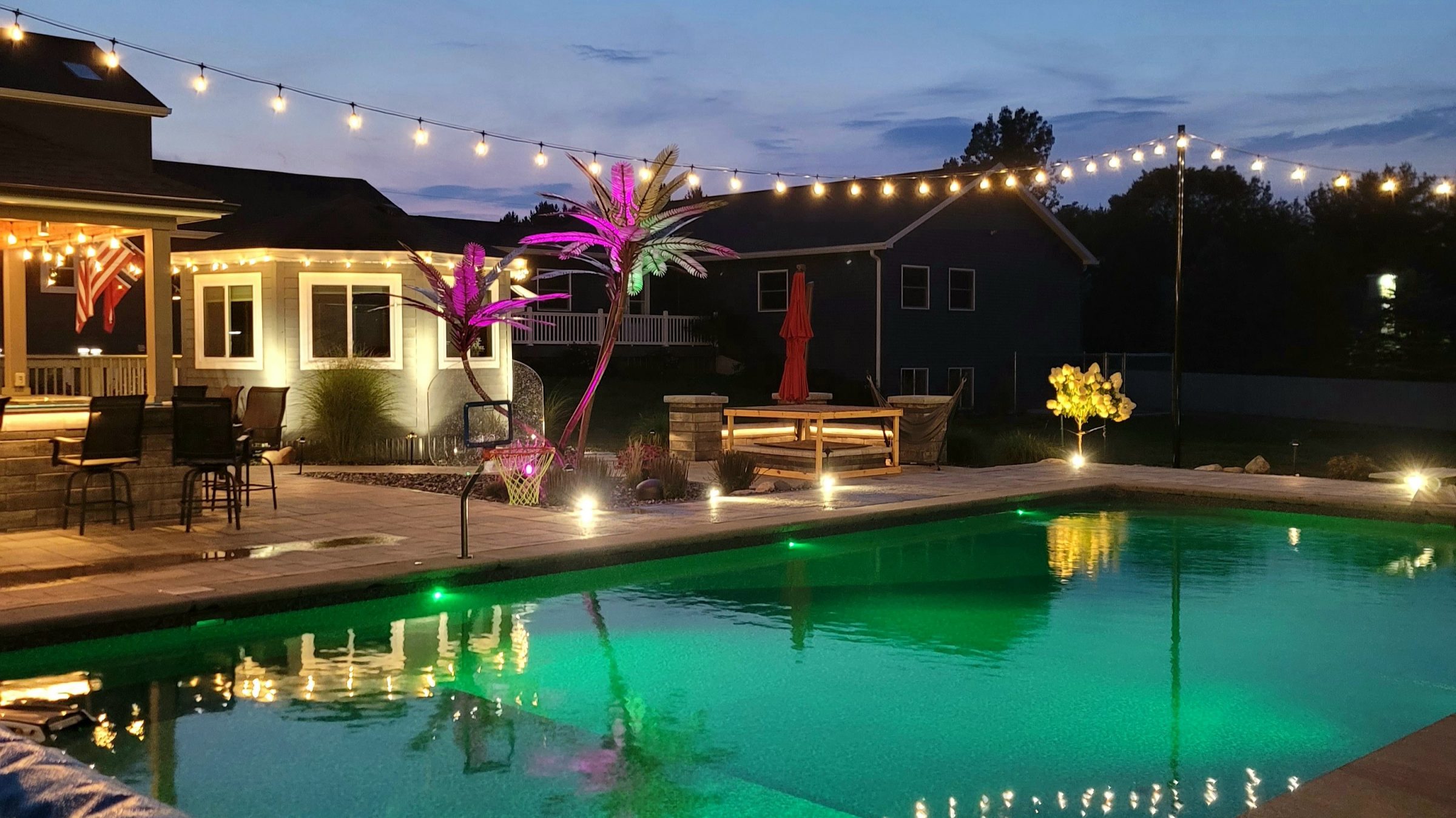 Illuminated backyard pool with string lights at dusk.