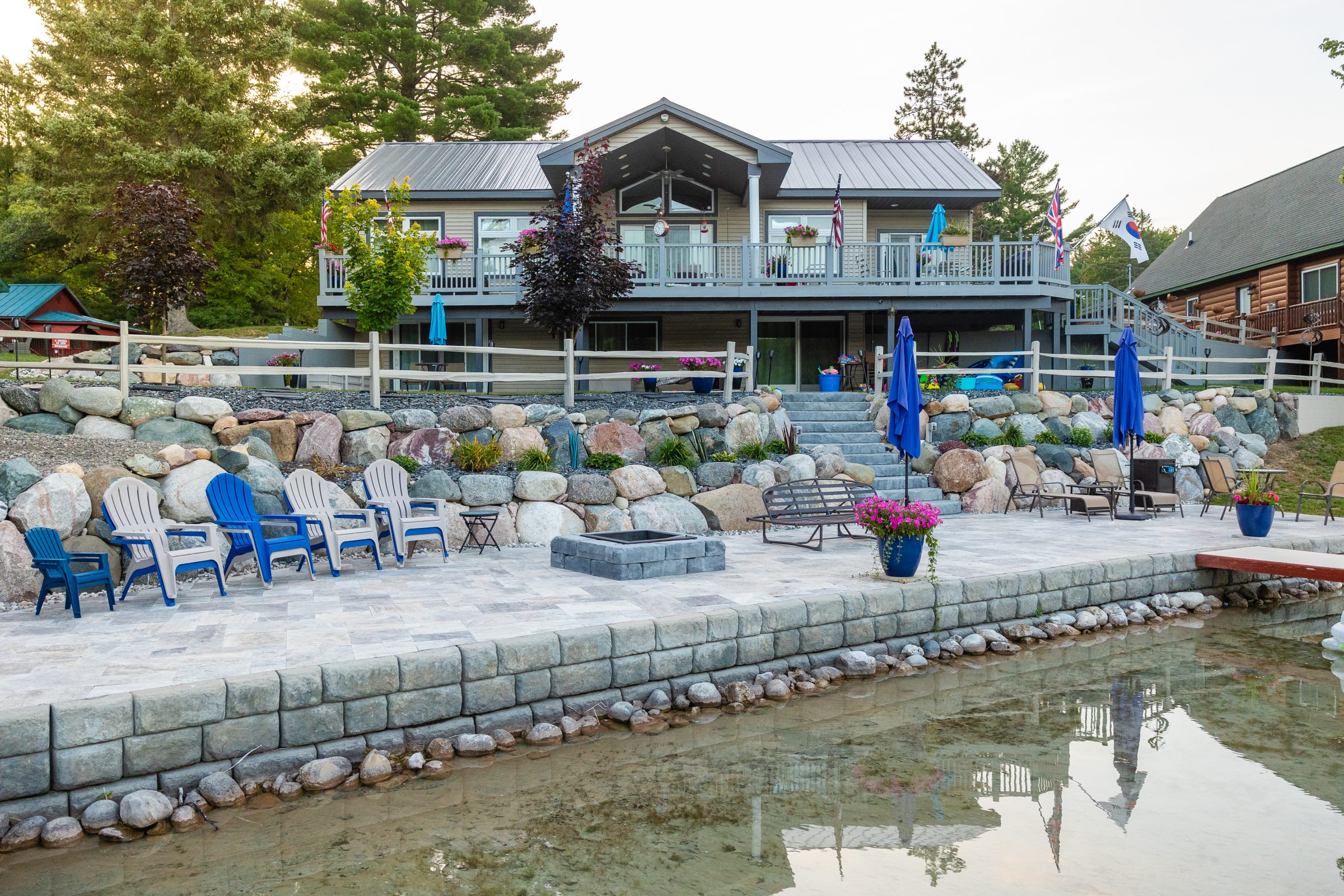 Lakeside home with patio and chairs, surrounded by trees.