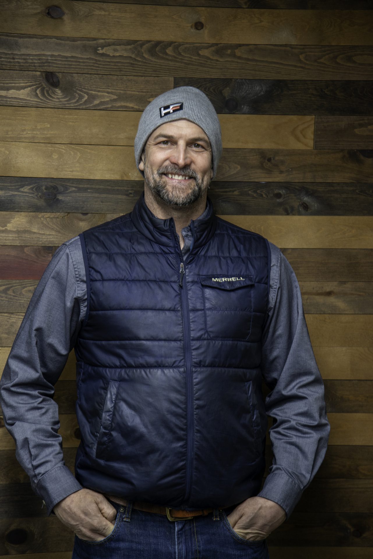 Smiling man in beanie and vest against wood backdrop.