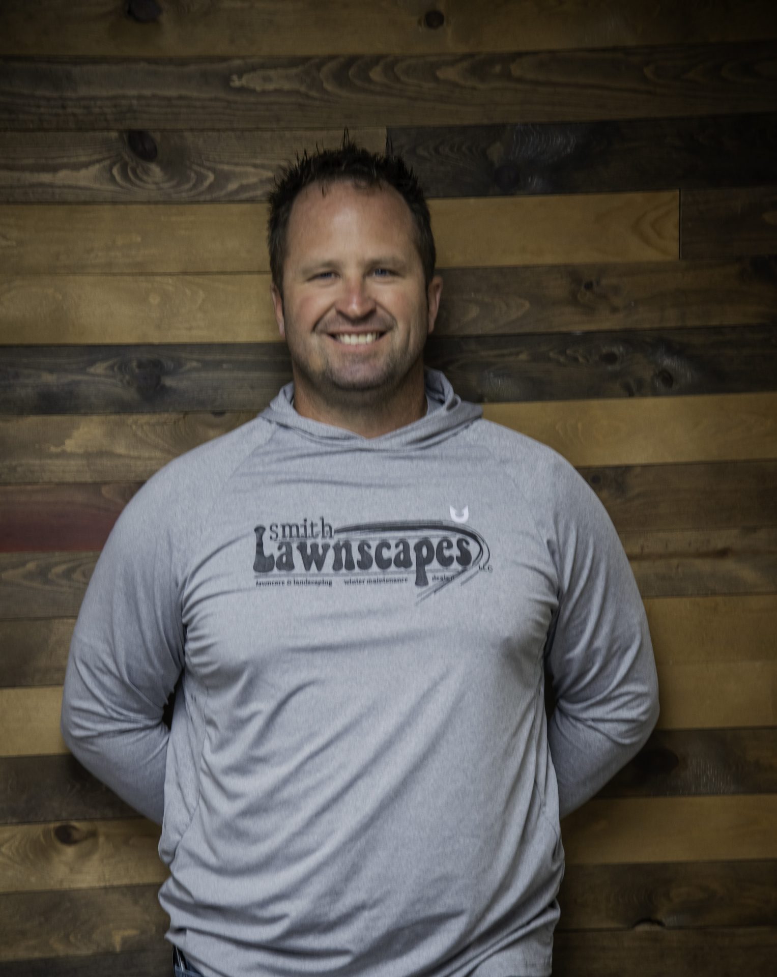 Person smiling in gray 'Lawnscapes' shirt, wood background.