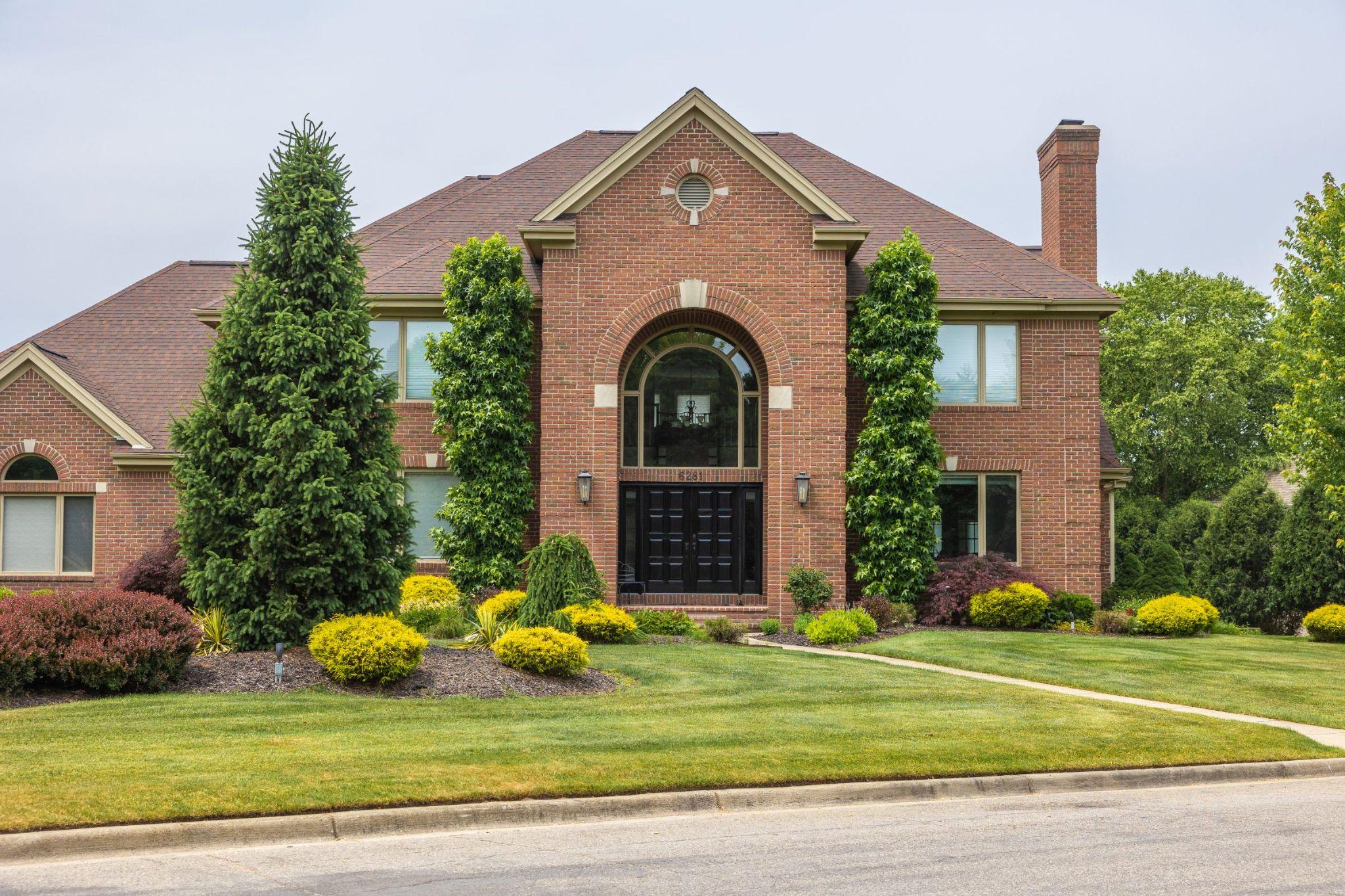 Brick home exterior with symmetrical landscaping, trimmed shrubs, mature trees, and a well-maintained front lawn.