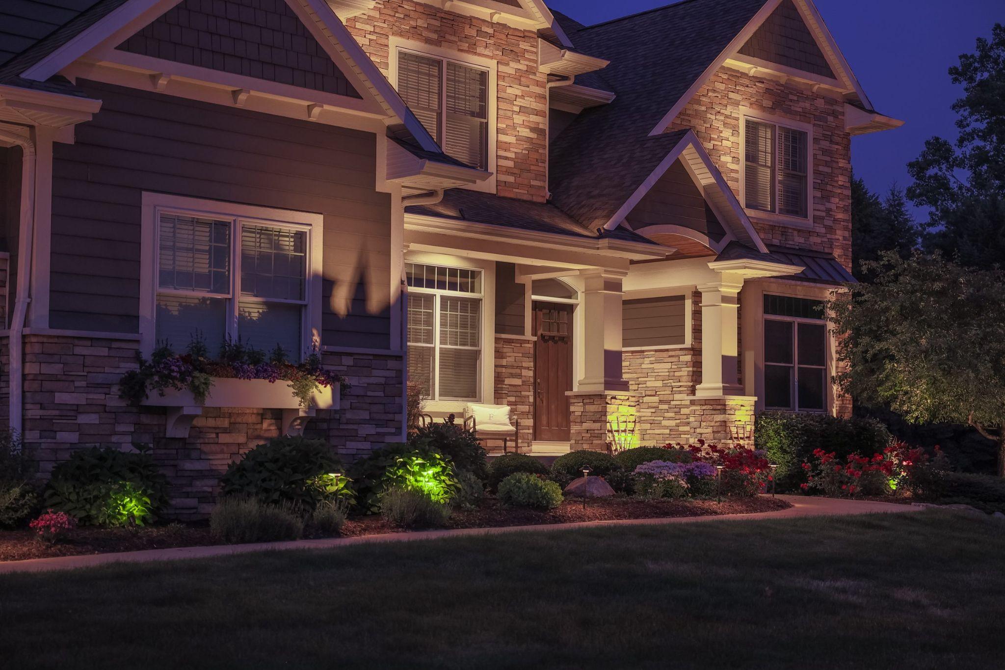 Evening view of a home exterior with landscape lighting highlighting stone accents, planting beds, and walkway.