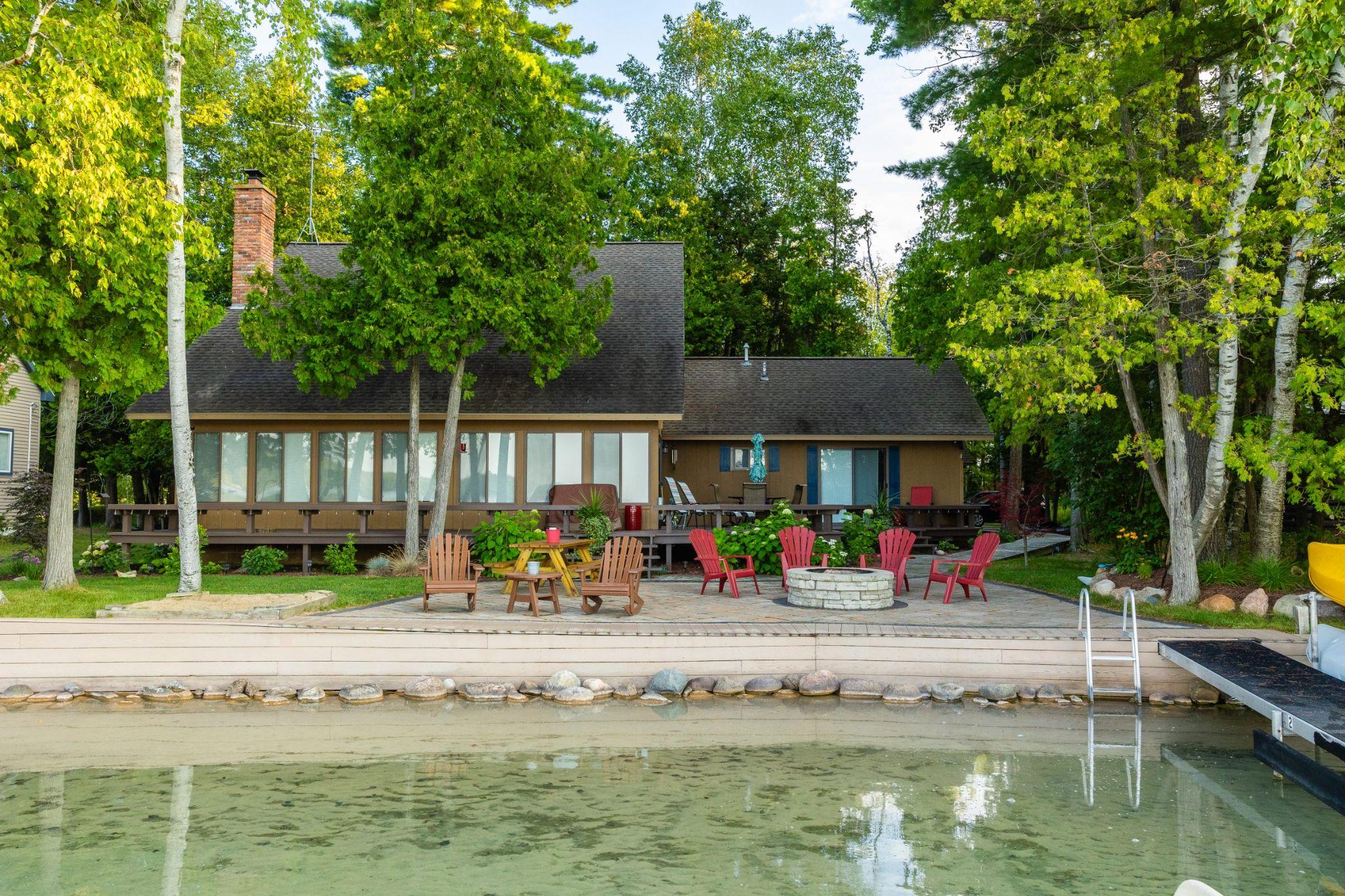 Lakeside patio with fire pit seating, Adirondack chairs, wooden deck, and calm water in the foreground.